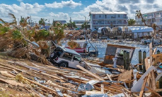 Area devastated by a hurricane shows damaged buildings & vehicles and downed trees and debris.
