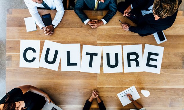 Six business people are shot from above as they sit around a table. Seven white pieces of paper are laid on the table in the middle of them, each with one letter on it. The papers together spell 