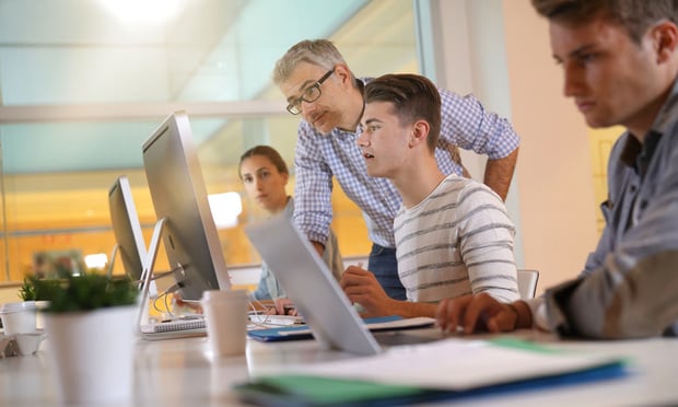 Three young people sit at computers while an older professional gives them instruction.