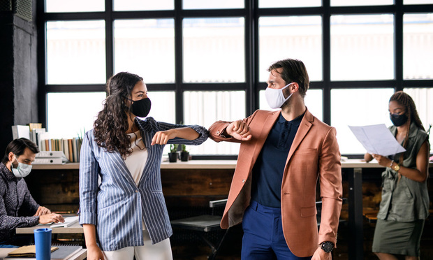Two young, masked professionals bump elbows in greeting at the office. 