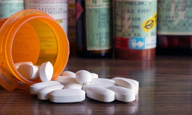 A bottle of white prescription pills is spilled on a table. A collection of other prescription bottles are in the background.