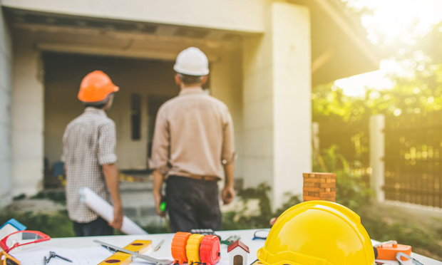 Two contractors stand with their back to the camera, looking at a house. There are various construction materials between them and the camera.