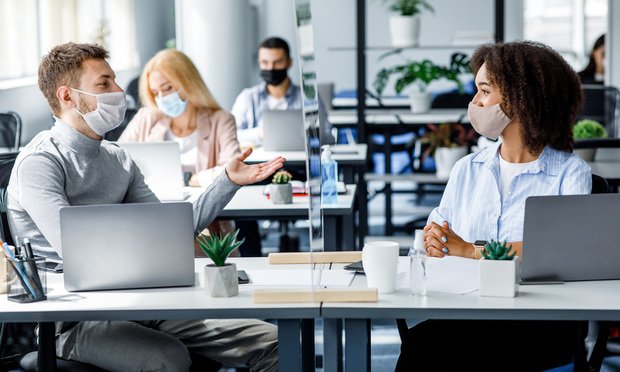 Two young professionals wearing masks sit at a desk talking.