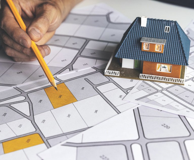 A small, model house sits on a map as a person points to a specific lot.