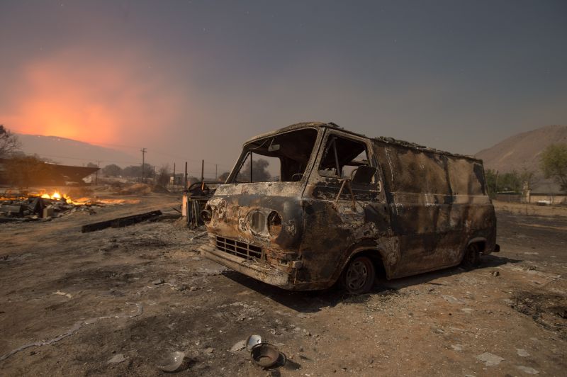 a burned van rests in a lot while a wildfire glows on the horizon