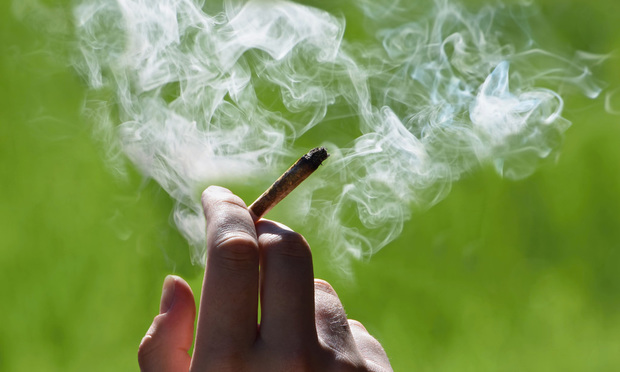 A hand holds a lit cannabis joint as smoke billows around. 