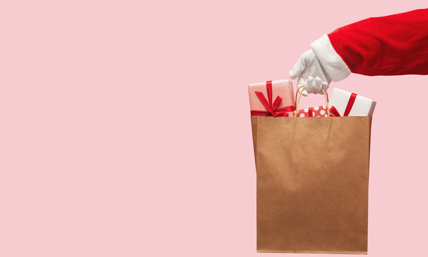 A Santa Claus hand holding a paper bag full of gifts in front of a pink background.