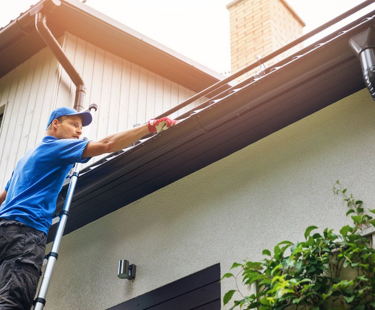 A man stands on a ladder, cleaning out a home's gutters.